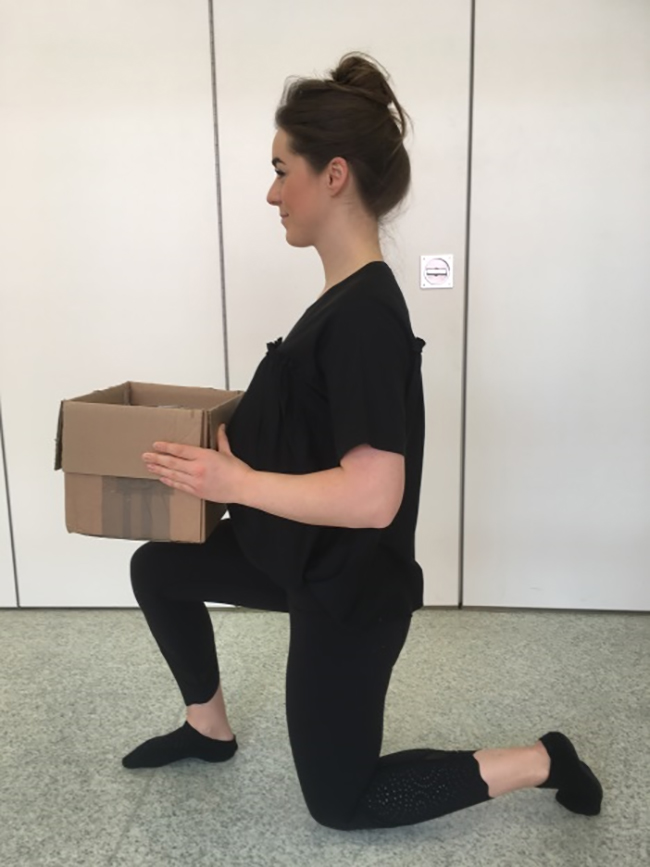 Woman bending down on one knee with her hands supporting a box on her raised bent knee to demonstrate safe lifting